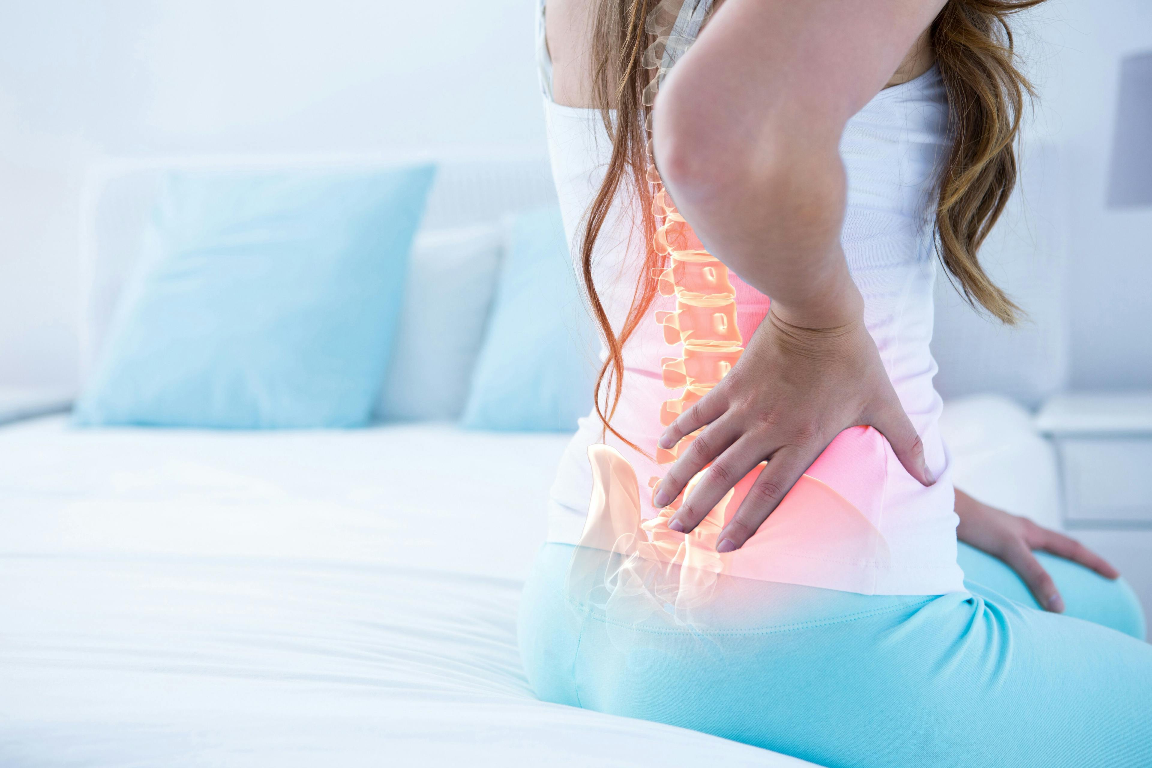 Woman sitting on bed with back pain illustrated by a highlighted spine | Image credit: WavebreakMediaMicro - stock.adobe.com